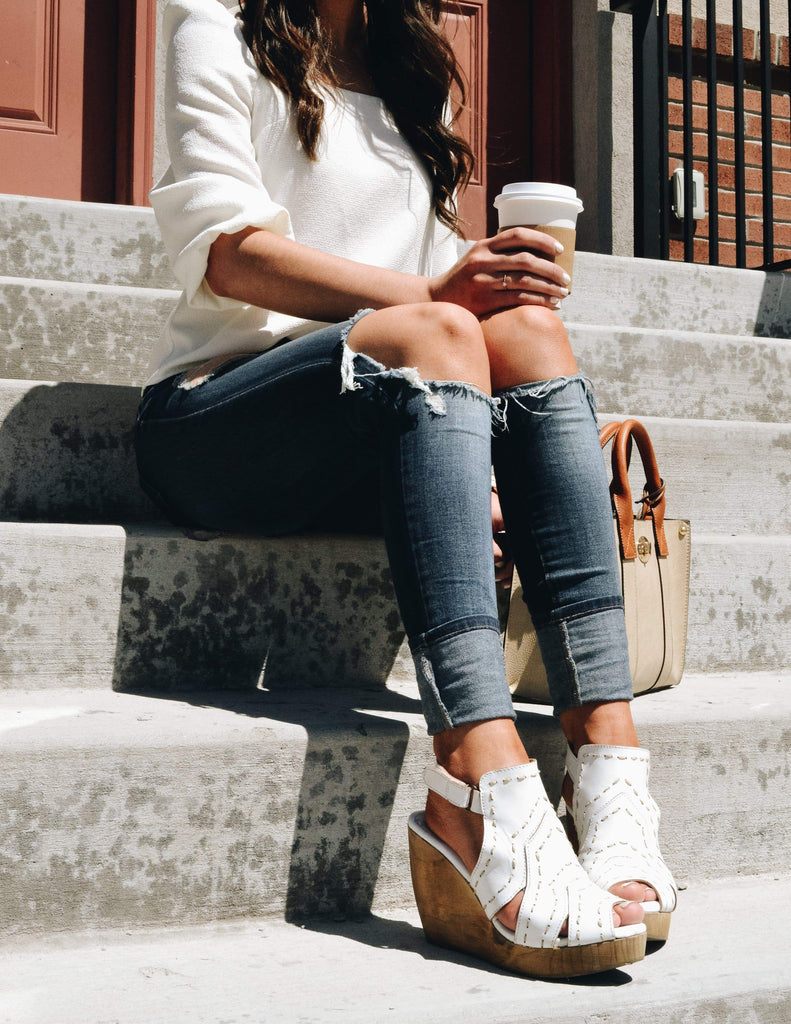 Model sitting on steps outside in white shirt, denim, and white wedges
