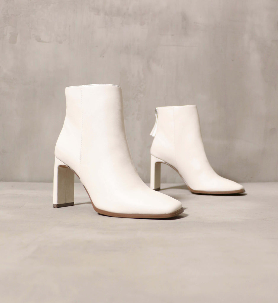 A white ankle bootie with a thin rectangular heel on a cement backdrop - Elle Bleu