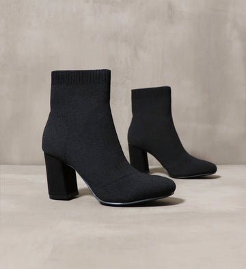 A pair of black knit stretchy ankle booties on a concrete background - Elle Bleu