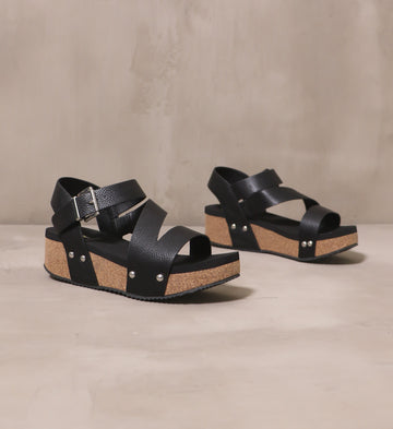 pair of all strapped in wedge with pebble leather black straps and cork soles on cement background