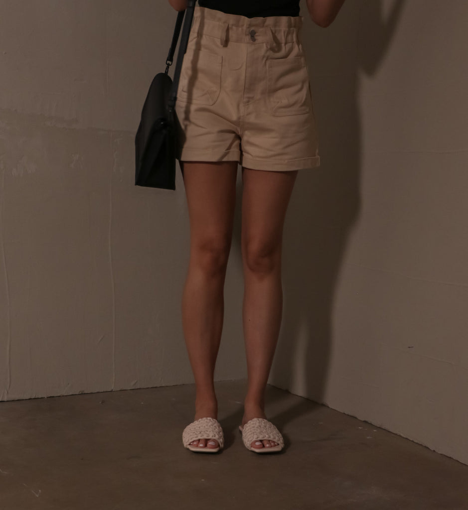 model standing in cement room wearing beige shorts and open toe macra-meet me at the dock slide sandals holding black purse