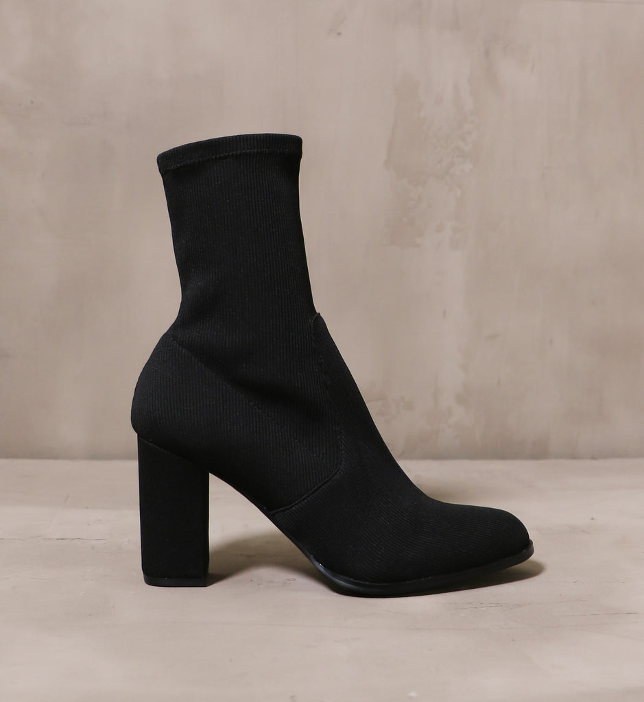outer side of the all black knit happens boot with fabric wrapped block heel on cement background