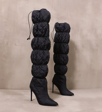 pair of all black mother puffer boots with banded tall shafts and stiletto heels on cement background