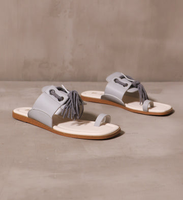 a pair of light grey blue tassel in the sky slide sandals with thin big toe strap and leather strap across the top of the shoe