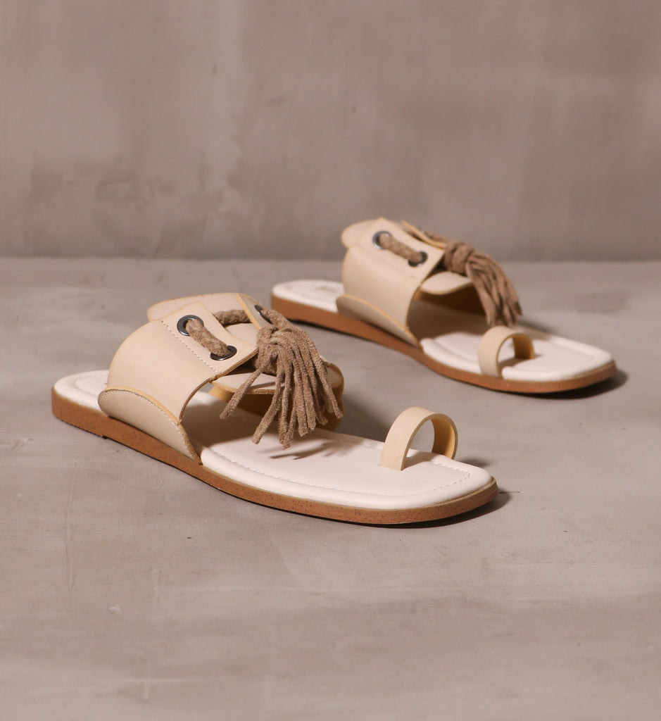 a pair of beige tassel in the sky slide with beige toe loop and strap across the top of the shoe on off white insole